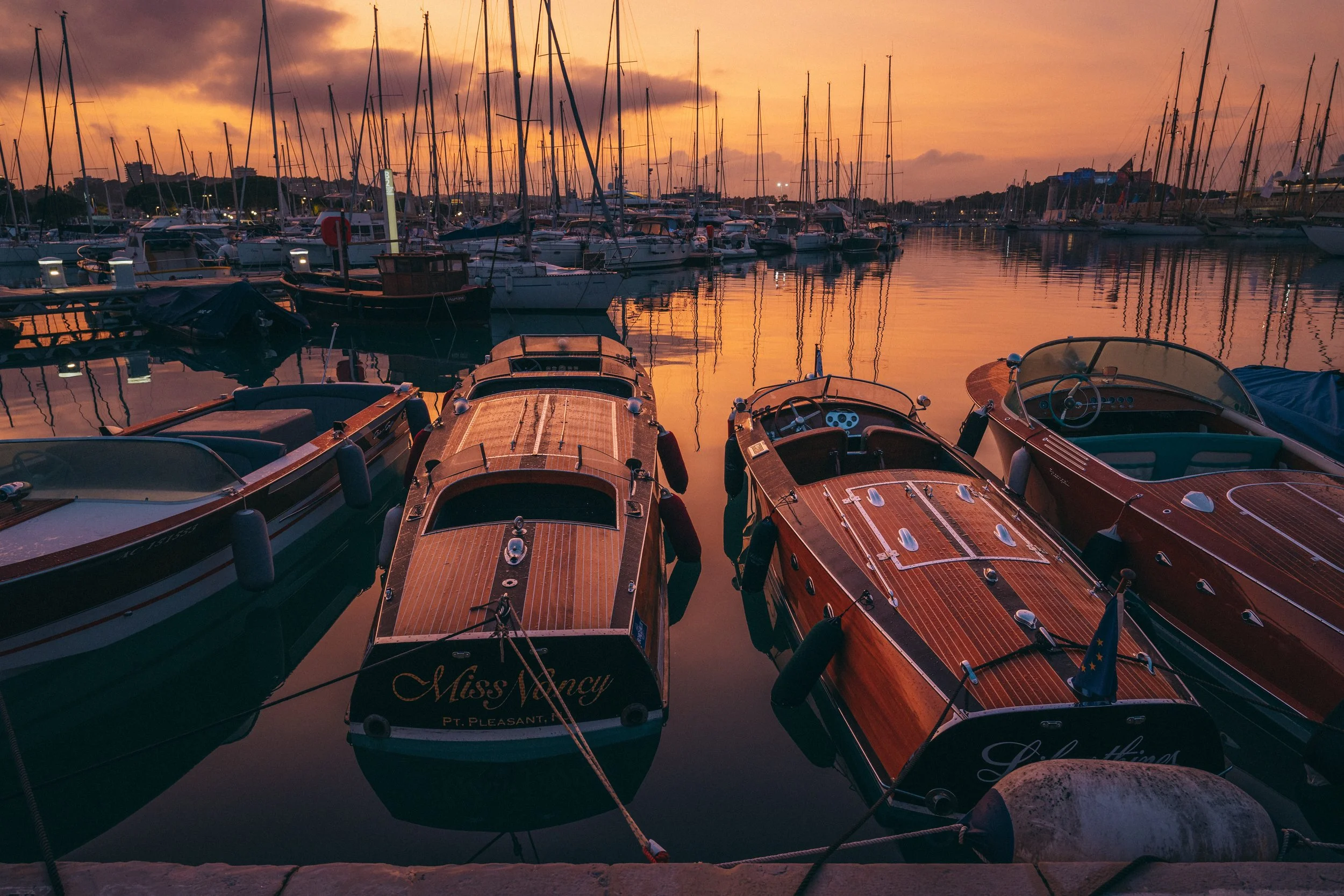 Bateaux à quai au coucher du soleil dans un port, avec un ciel orange et des eaux calmes qui reflètent les voiliers.