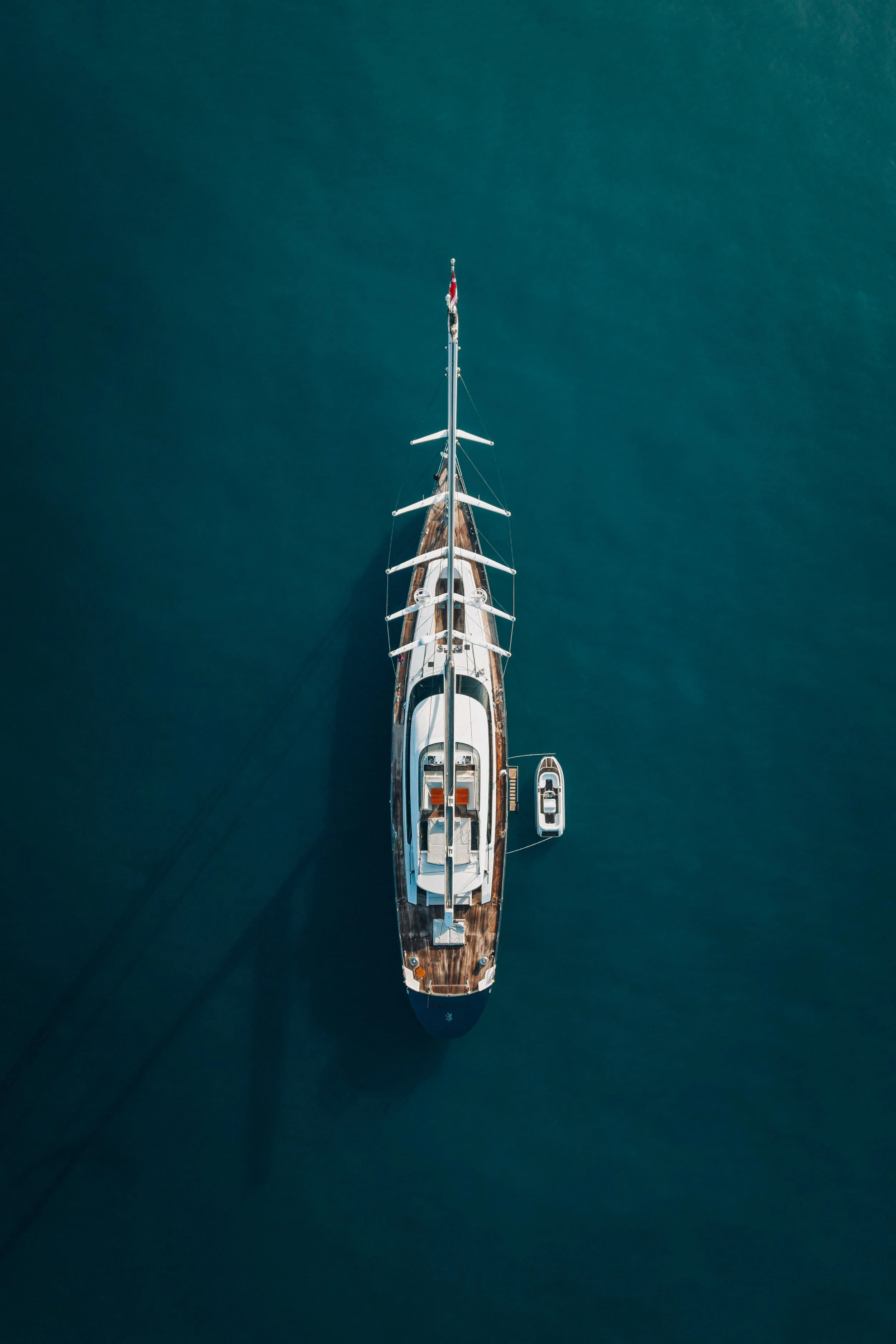 Vue aérienne d'un yacht en mer avec une coque en bois et un mât. Le bateau est vu du dessus, flottant sur une eau bleue calme.