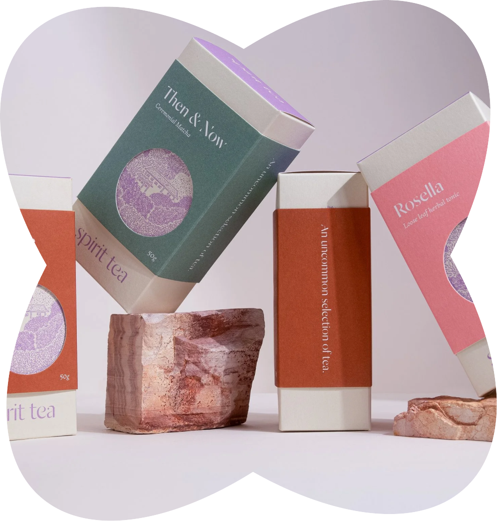 Boxes of herbal tea labeled "Spirit Tea," "Then & Now Ceremonial Matcha," and "Rosella Loose leaf herbal tonic" arranged on rocks with a white background.