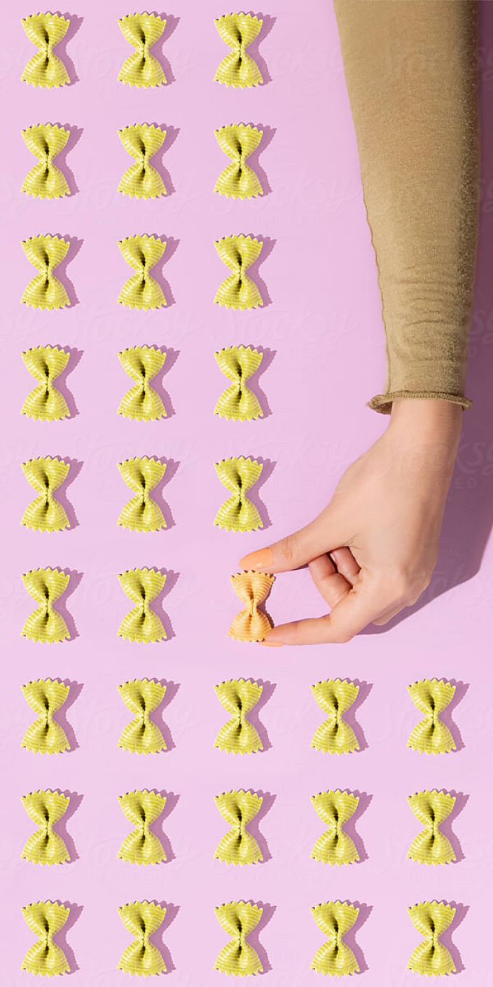 A person's hand is picking up an orange-colored piece of fusilli pasta from a pink surface. There are multiple rows of yellow fusilli pasta arranged on the surface.