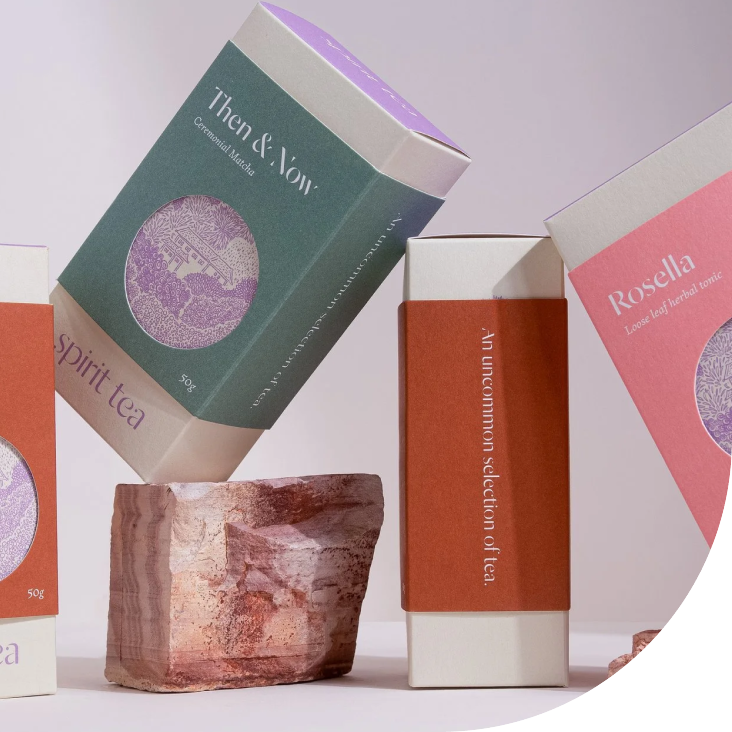 Display of tea packages, labeled "Then & Now" and "Rosella," along with a pinkish rock in the foreground.