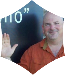 A smiling man in an orange shirt standing next to a dark surface with partially visible text.