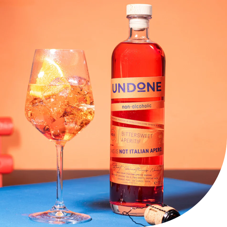 A wine glass filled with a pink, fizzy beverage with ice and lemon slices next to a tall, cylindrical bottle labeled "UNDONE non-alcoholic" and "Bittersweet Aperitif" on a blue surface against a peach-colored background.
