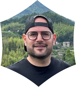 A man with glasses and a black baseball cap turned backwards, smiling outdoors with a mountain and greenery in the background.