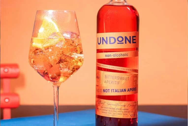 A glass of pinkish-colored tonic water with ice and lemon slices next to a bottle labeled 'UNDONE' which is a non-alcoholic beverage, flavored as bittersweet aperitif.