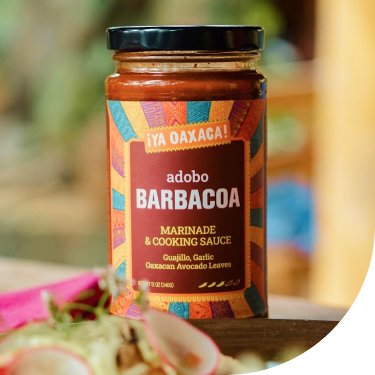 A jar of Adobo Barbacoa marinade and cooking sauce with a colorful label that includes text in both Spanish and English, placed on a wooden surface with some blurred food in the foreground.