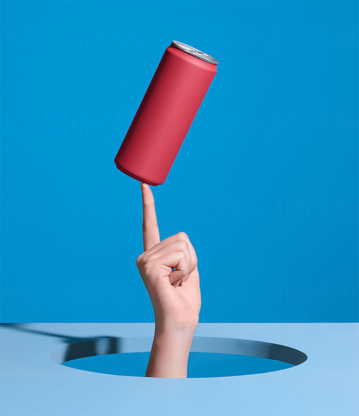 A hand with the index finger pointing up, emerging from a hole in a blue surface, balancing a red soda can against a blue background.