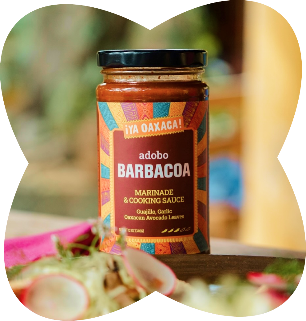 A jar of Adobo Barbacoa marinade and cooking sauce with flavors of guajillo, garlic, and Oaxacan avocado leaves, placed on a wooden surface with blurred flowers in the foreground.
