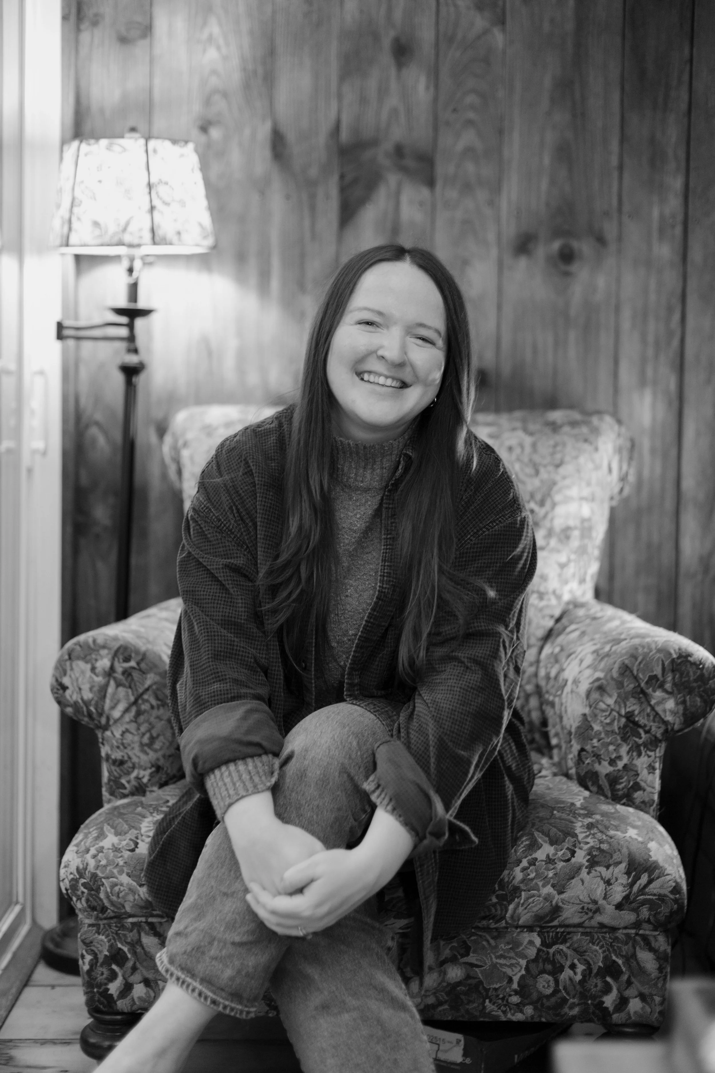 A smiling woman with long hair sitting on a floral upholstered armchair in a room with wooden walls, beside a lit floor lamp.