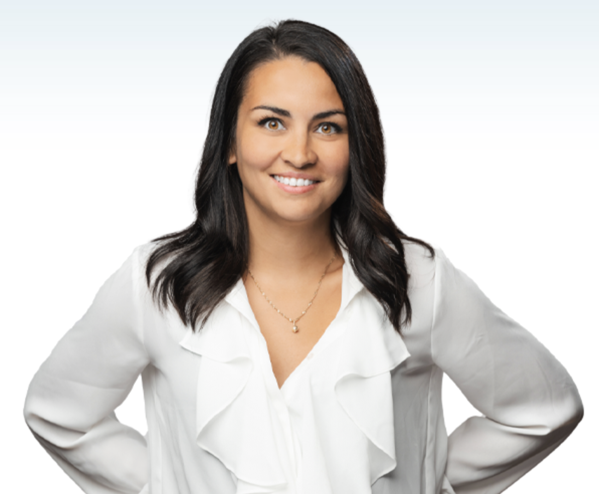 A woman with dark brown, wavy hair wearing a white blouse and a gold necklace, smiling with her hands on her hips.