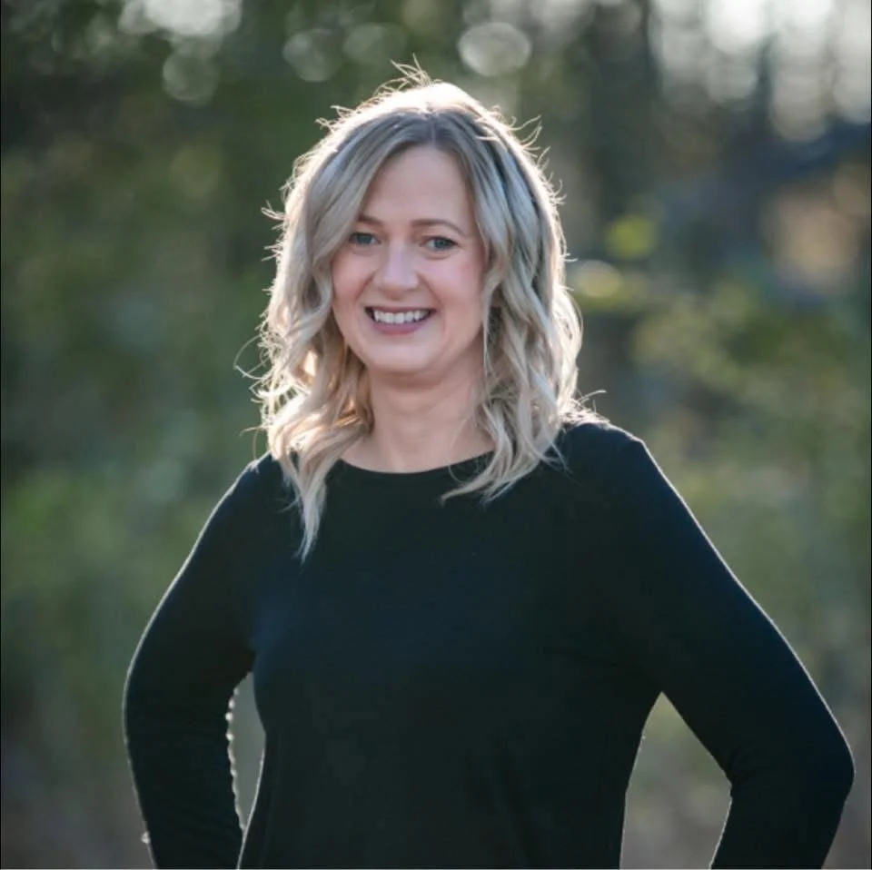 A woman with wavy blonde hair smiling outdoors with trees in the background.