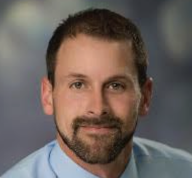 A man with short dark hair, a beard, and mustache, wearing a light blue shirt, looking at the camera with a slight smile.