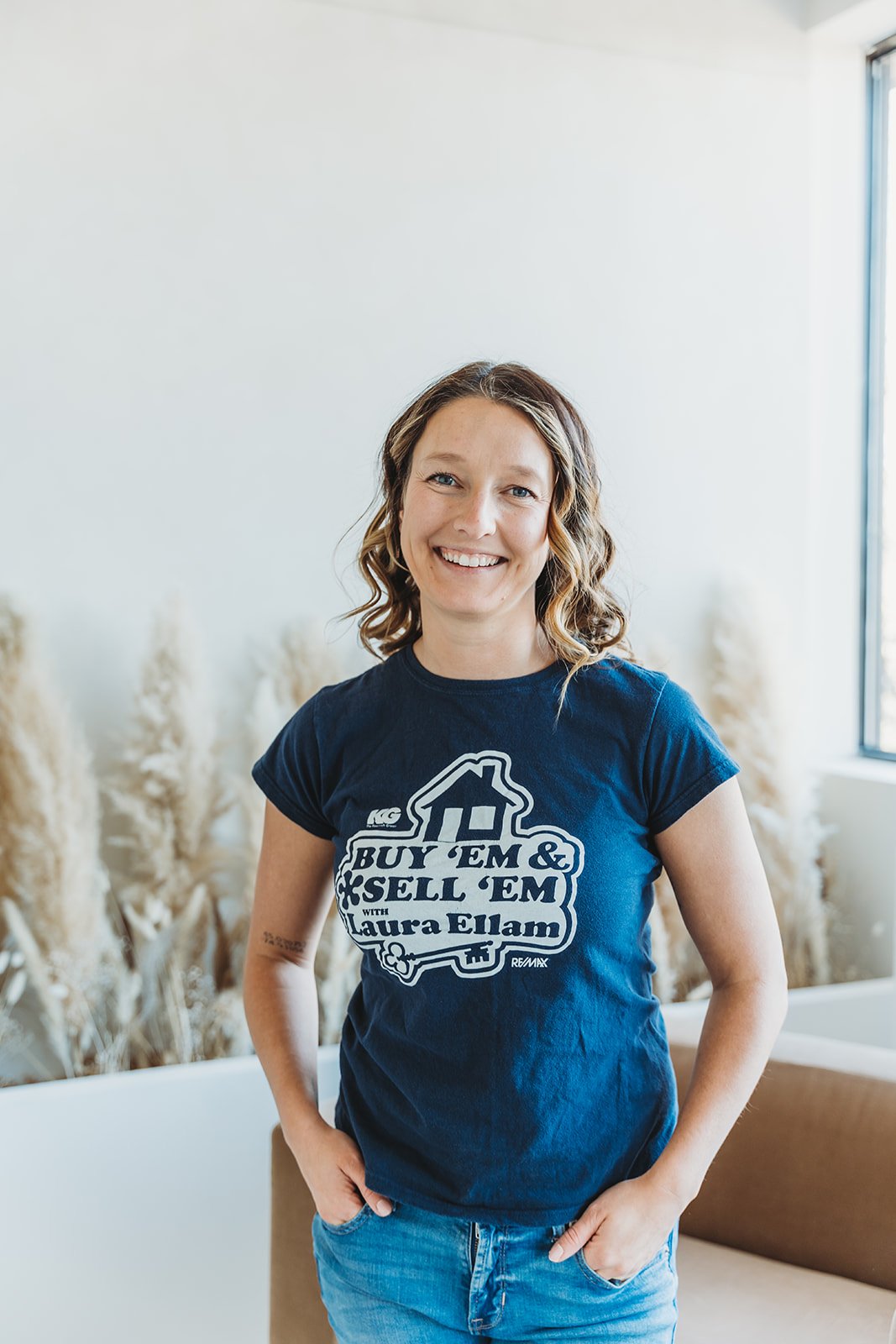 Woman with shoulder-length curly hair smiling, wearing a navy t-shirt that says 'Buy 'Em & Sell 'Em with Laura Ellam,' standing in a room with light-colored walls and dried decorative plants.