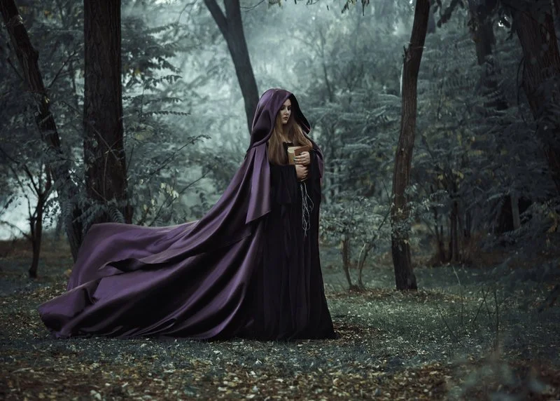 A woman dressed as a wizard or sorceress in a purple cloak, holding a cup, standing in a forest with dense trees and misty atmosphere.