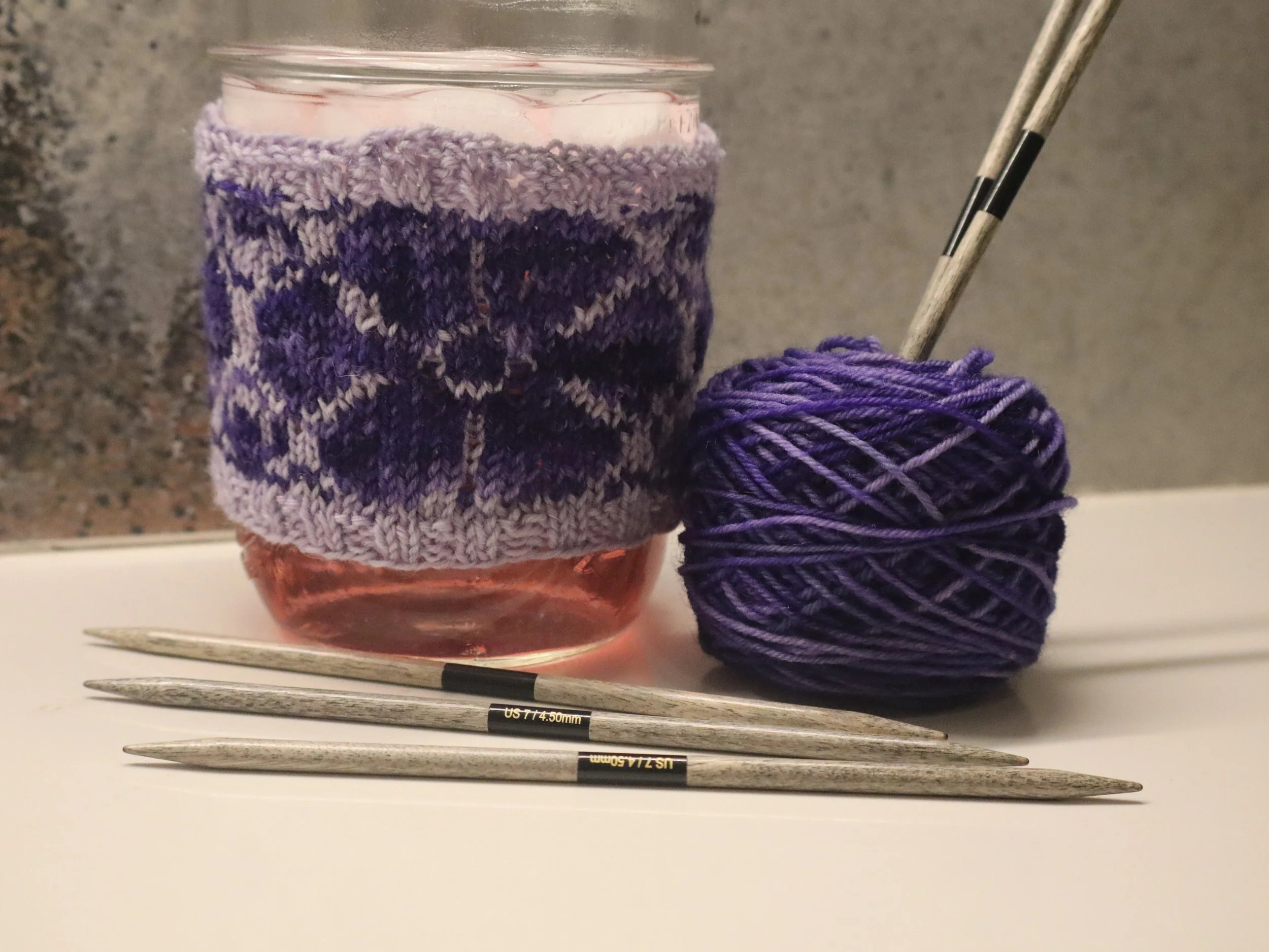Hand knit jar koozie with yarn dyed by Fox & Owl Dye Lab shown with knitting needles.