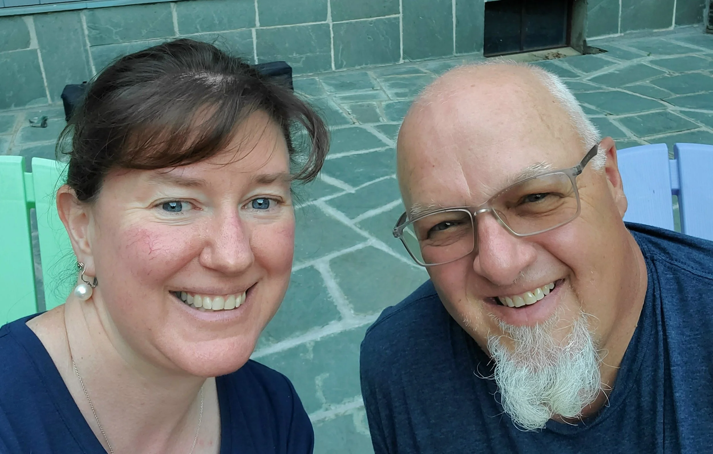 A smiling woman with short brown hair, blue eyes, wearing pearl earrings and a navy blue top, taking a selfie with a smiling man with a white beard, glasses, and a dark shirt, outdoors on a tiled patio with colorful chairs in the background.