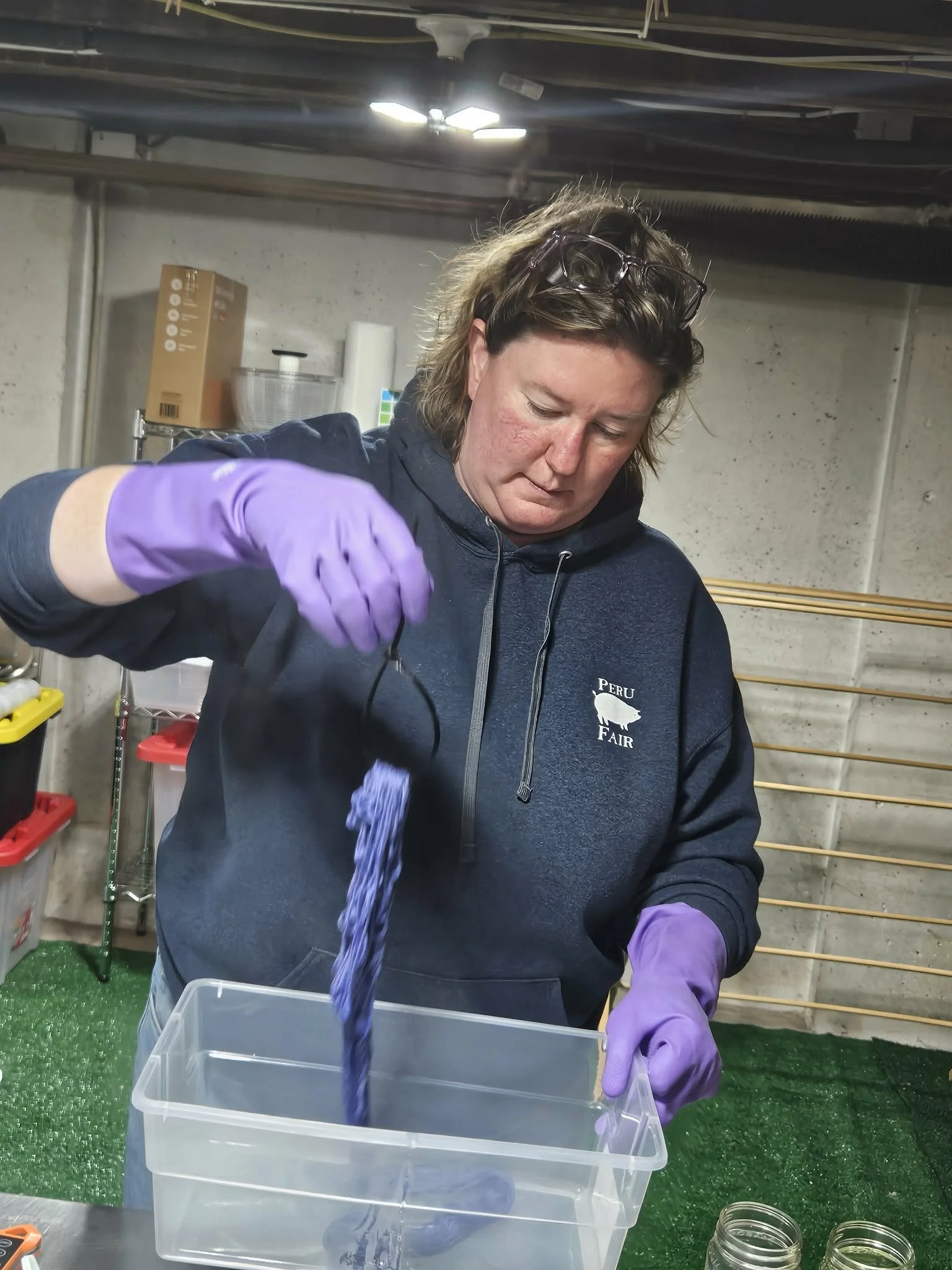 A woman with curly hair wearing glasses on her head and purple gloves is pouring purple liquid into a clear plastic container. The setting is a dye lab  with shelves and equipment in the background.