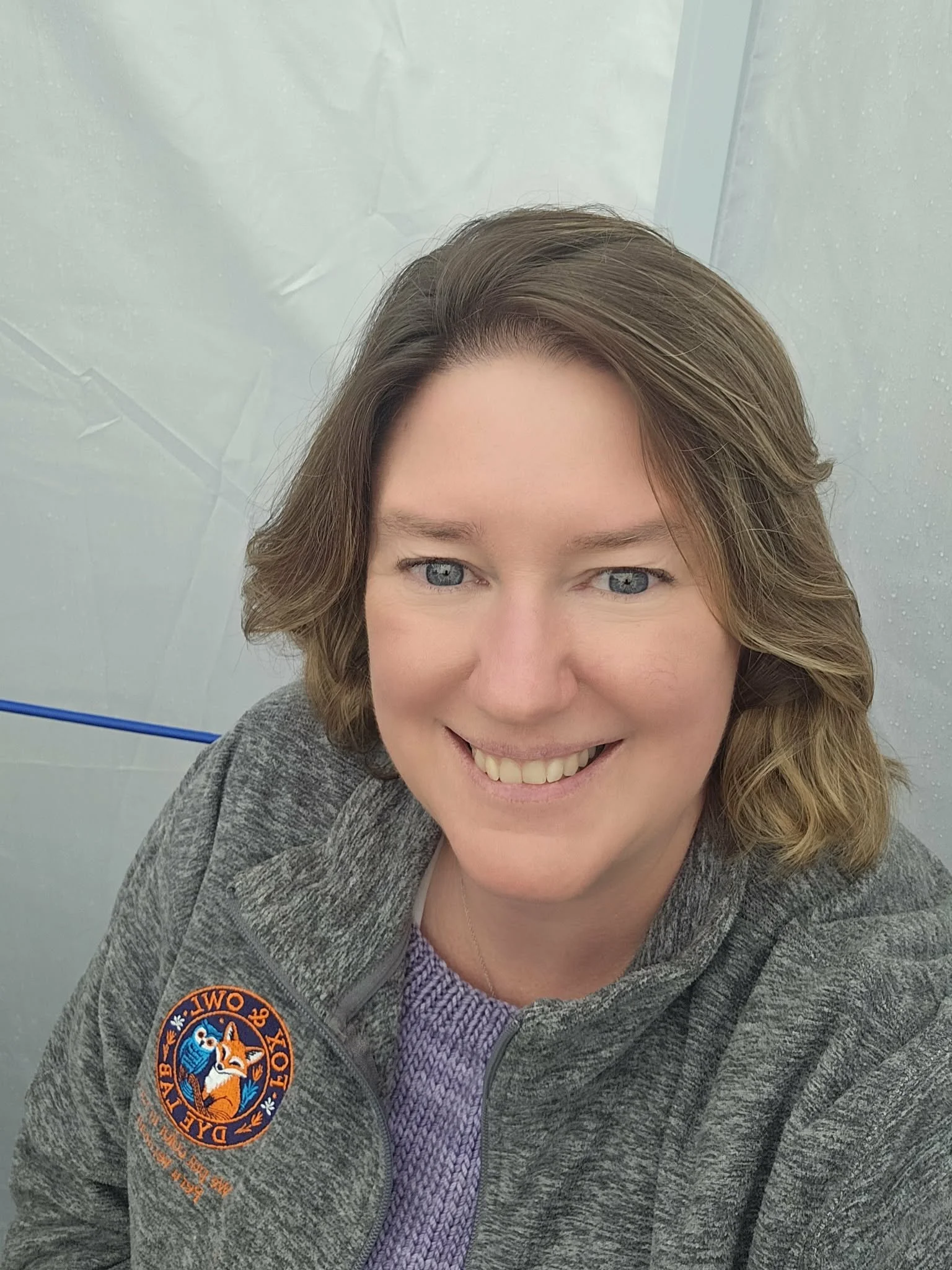 Close-up of a woman with light brown, wavy hair, smiling, wearing a gray jacket with a round patch on the sleeve, gray sleeve, and a purple sweater underneath, against a light-colored background.