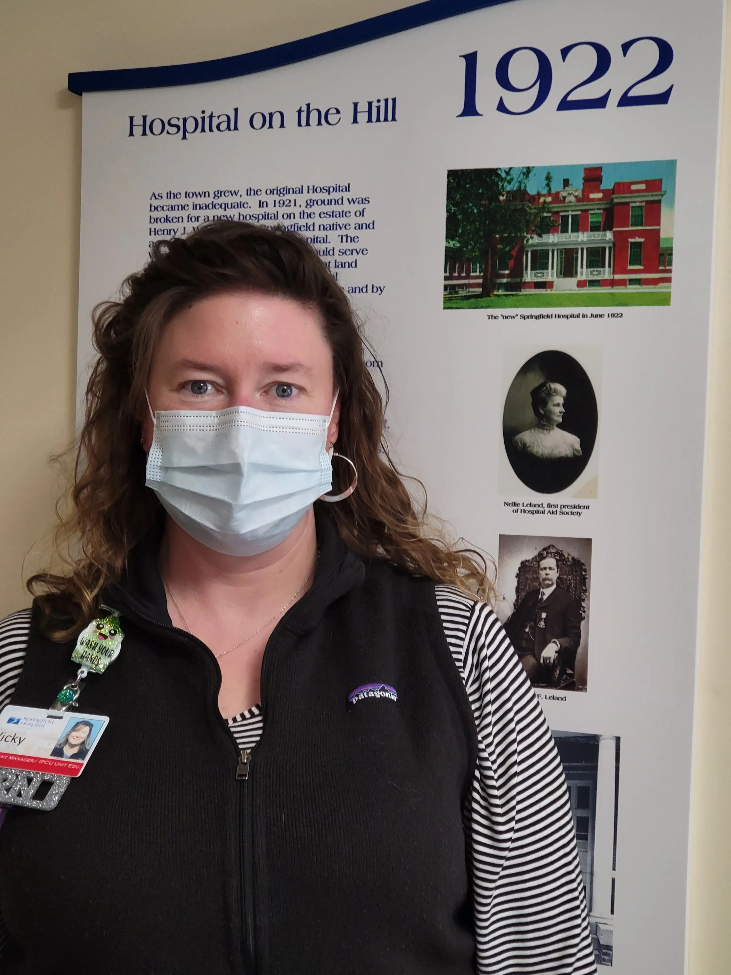 A woman wearing a face mask and a black vest with a name badge, standing in front of an informational poster about the history of Springfield Hospital, dated June 1922.