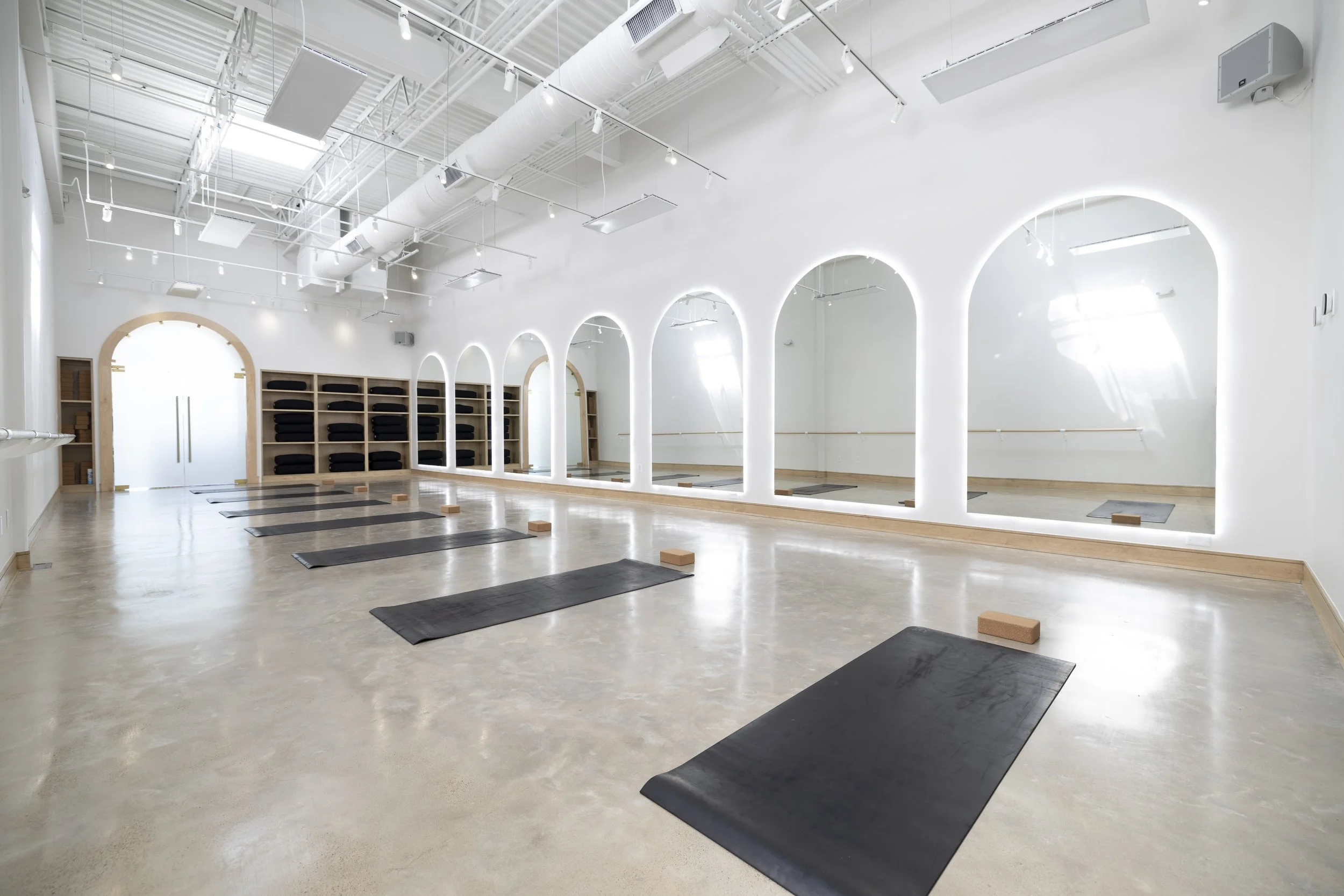 Empty yoga studio with wooden floors, black mats, and yoga blocks, large windows with blinds, and ballet barre on the wall.