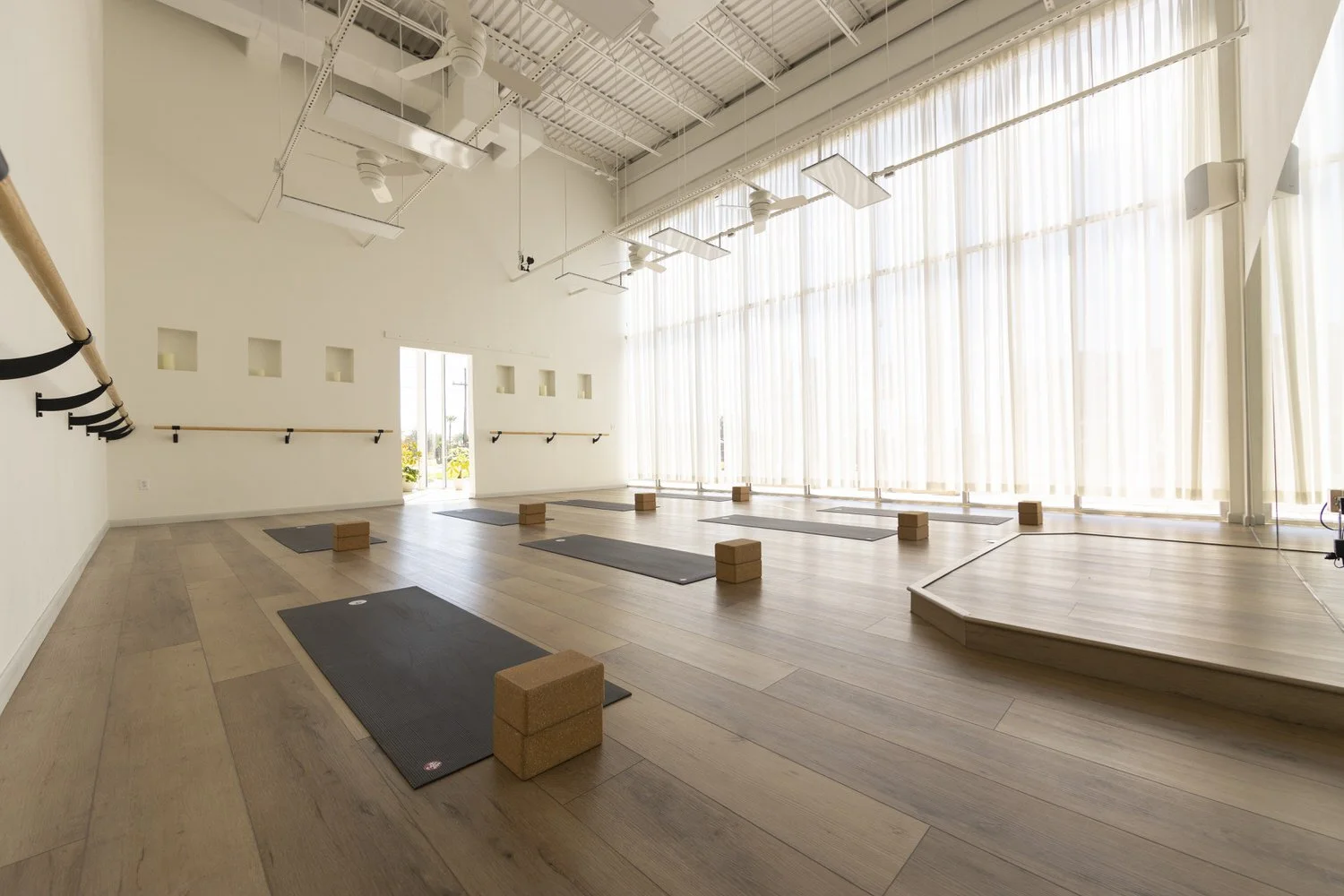 An empty yoga or fitness studio with wooden floors, large windows with curtains, black mats on the floor, wooden blocks, and wall-mounted ballet barres.