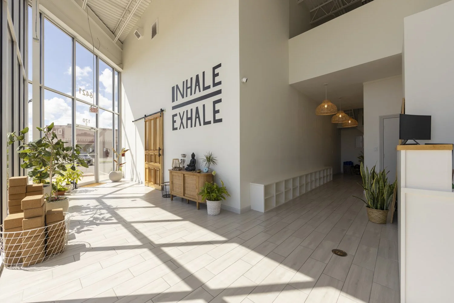 Bright, spacious yoga studio with large floor-to-ceiling windows, natural sunlight, and plants. The wall has a motivational quote 'Inhale Exhale' in large black letters. Decor includes a wooden door, a side table with a Buddha statue and plants, and 