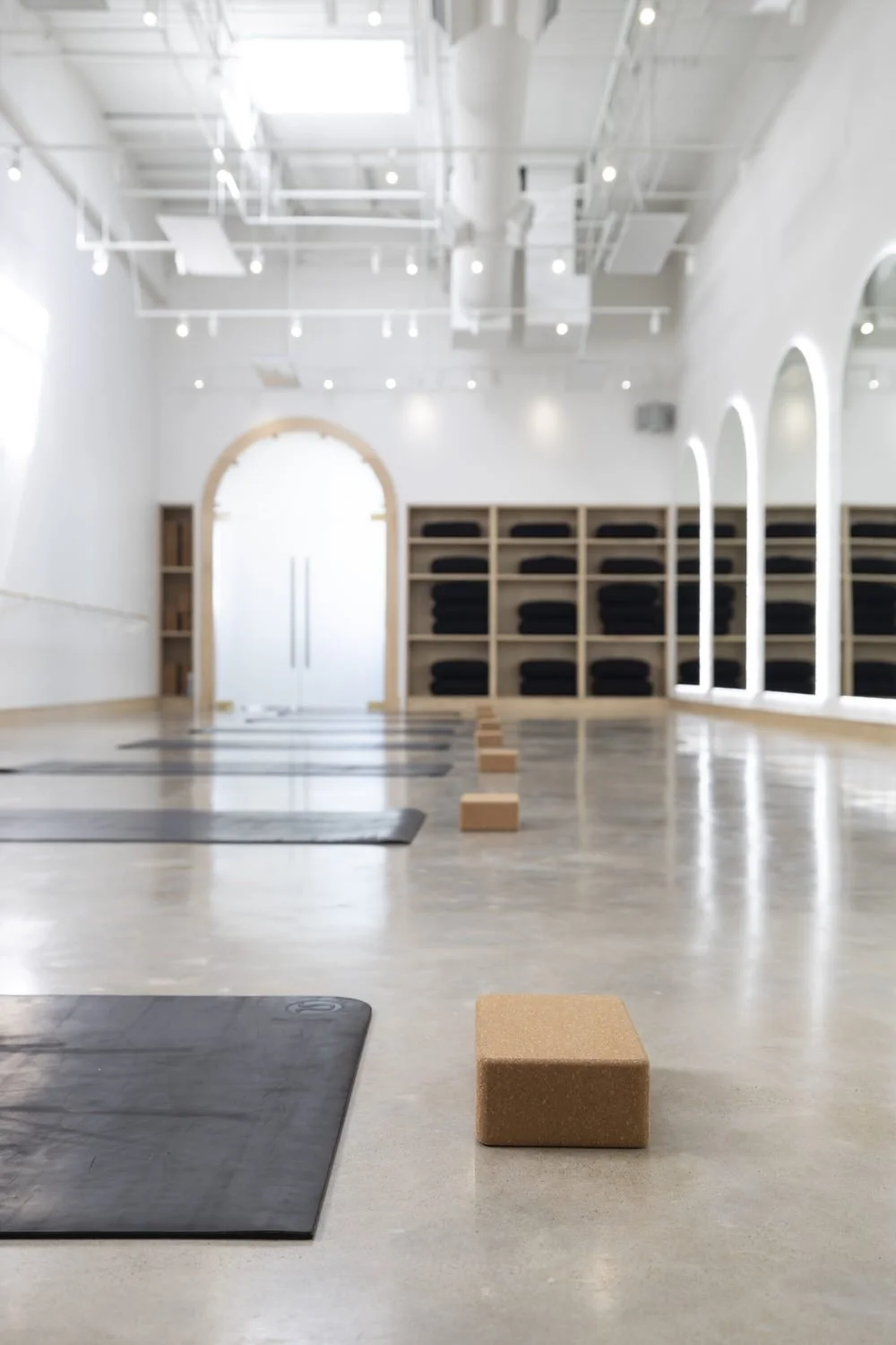 Yoga mats and cork blocks arranged in a studio with natural lighting, shelves with folded black towels, and large windows with arches.
