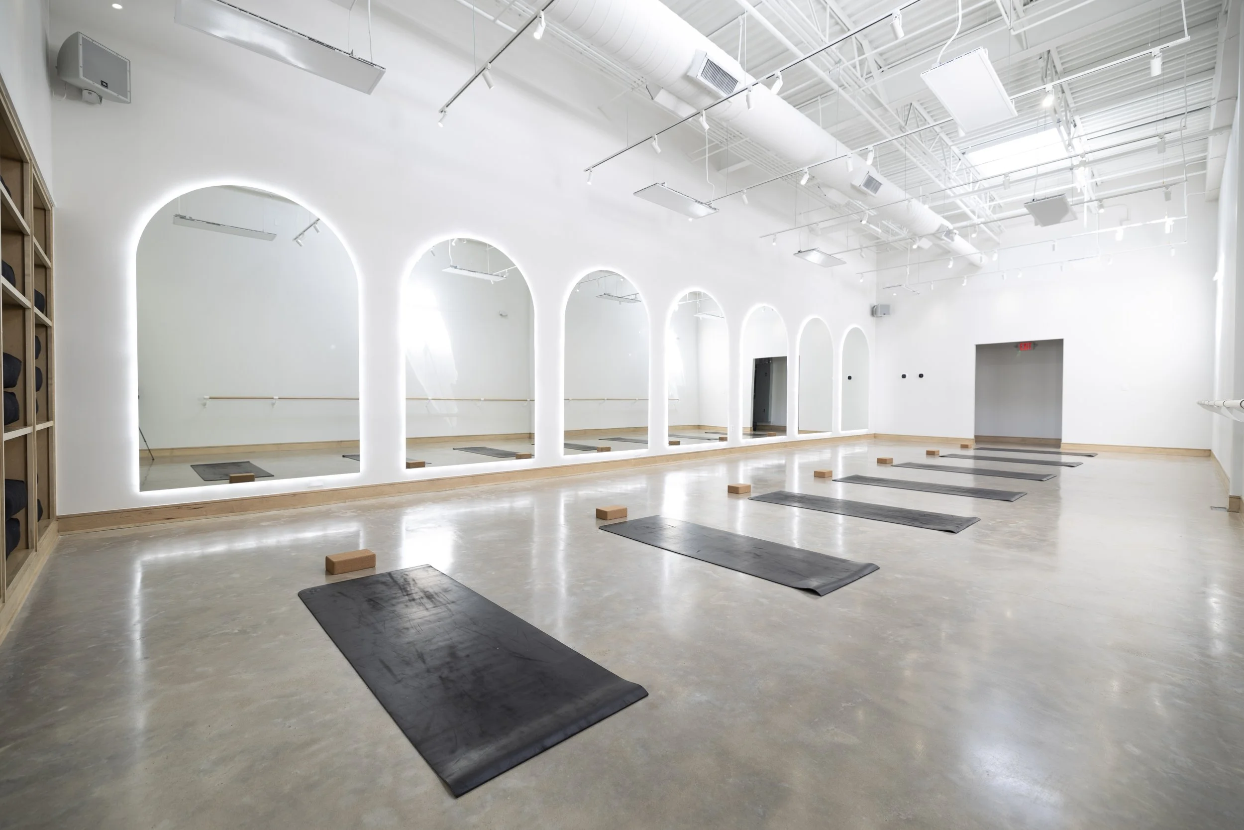 Empty yoga or fitness studio with black mats, wooden blocks, mirrors, white walls, and large arched windows in the background.