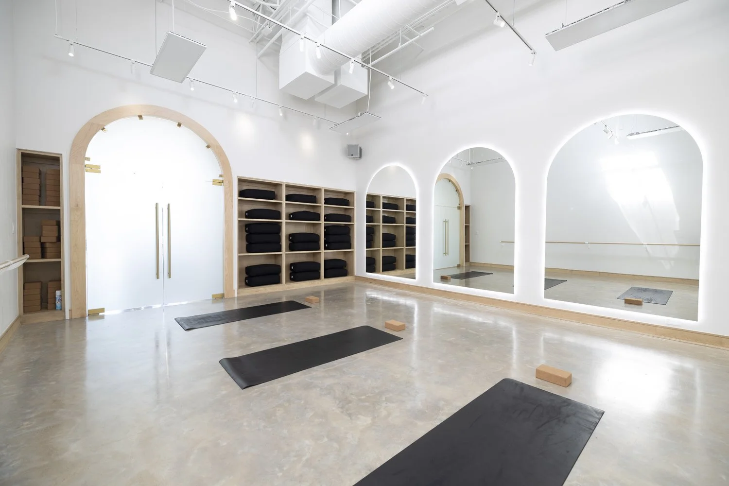 A bright, modern yoga or fitness studio with mirrors, black mats, and wooden shelves holding folded black blankets. The room has white walls, wooden accents, and a polished concrete floor.