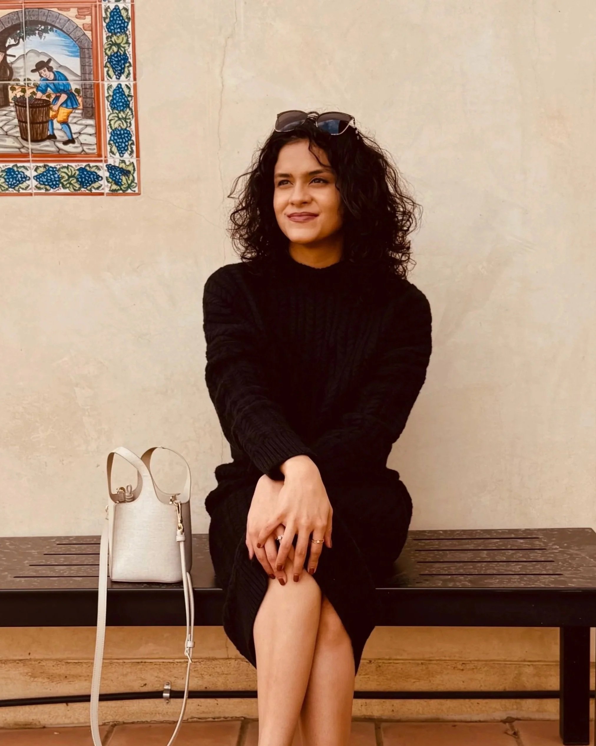 A woman with curly dark hair wearing a black sweater dress sits on a bench with her legs crossed and hands resting on her knees. A white handbag is placed beside her on the bench, and she has sunglasses on her head. The background includes a wall with a decorative tile picture of a person harvesting grapes.