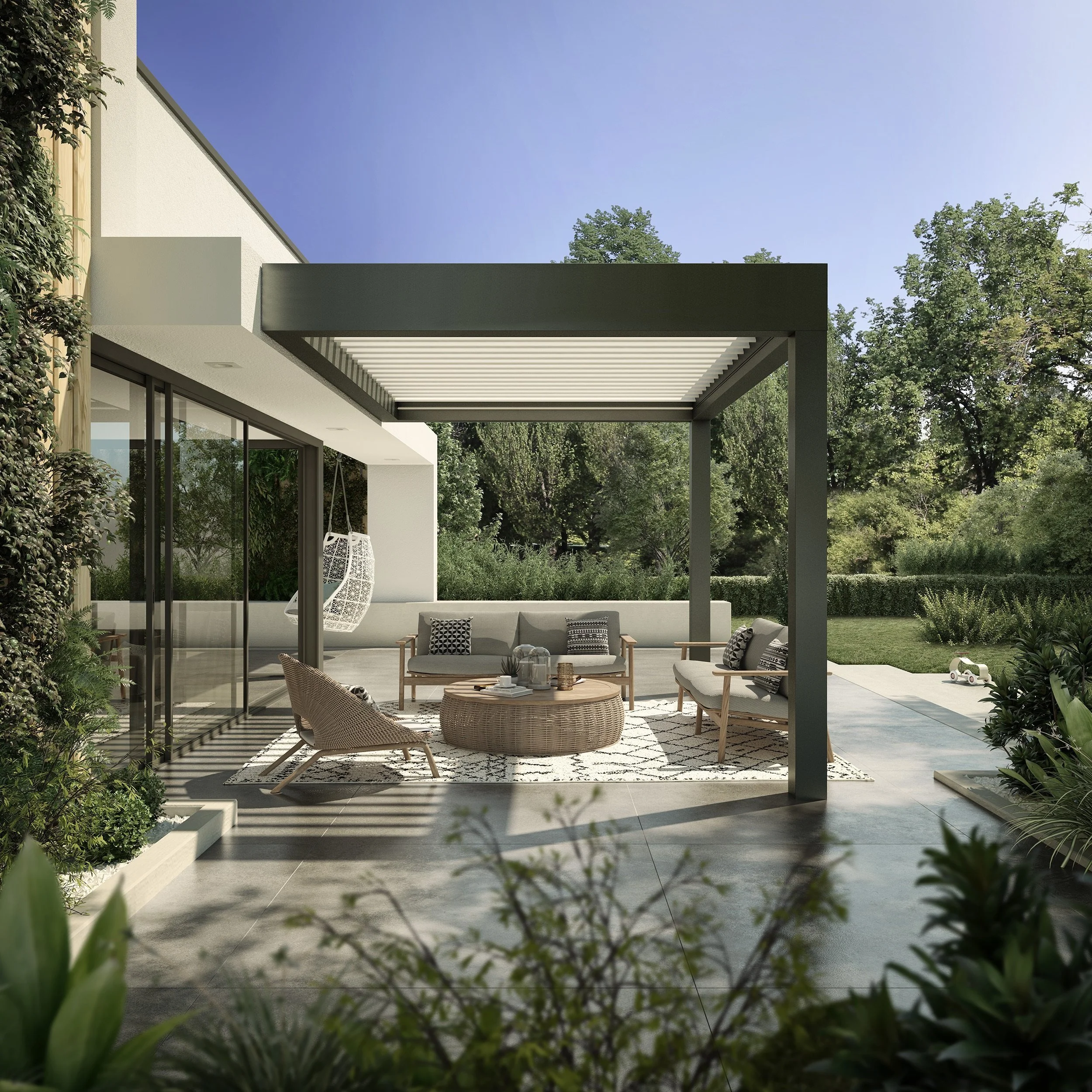 Outdoor patio with a pergola, modern furniture including sofas and chairs, displayed with decorative pillows, a round wicker coffee table with items on it, surrounded by greenery and trees, under a clear blue sky.