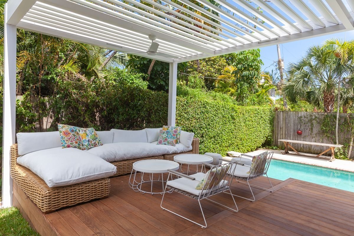 Outdoor patio with white cushioned wicker sectional sofa, floral throw pillows, white metal chairs with cushions, small round tables, overlooking a pool surrounded by green trees and a wooden fence, under a white pergola.