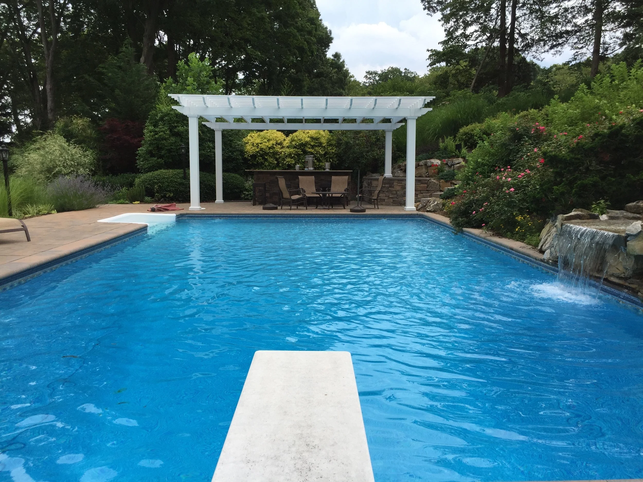 A backyard swimming pool with blue water, a diving board at the near end, and a waterfall feature on the right side. In the background, there is a pergola with outdoor furniture and a stone patio surrounded by lush green trees and bushes.