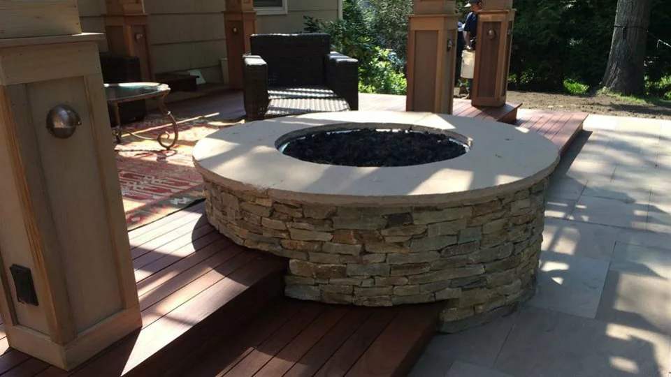 A circular stone fire pit with a concrete top on an outdoor patio, surrounded by wooden decking and some furniture.