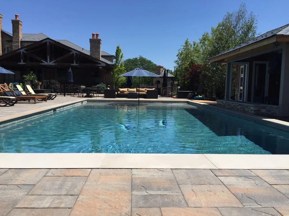 A rectangular backyard swimming pool with clear blue water, surrounded by a stone deck with lounge chairs, umbrellas, and outdoor furniture, with a house and trees in the background under a bright blue sky.