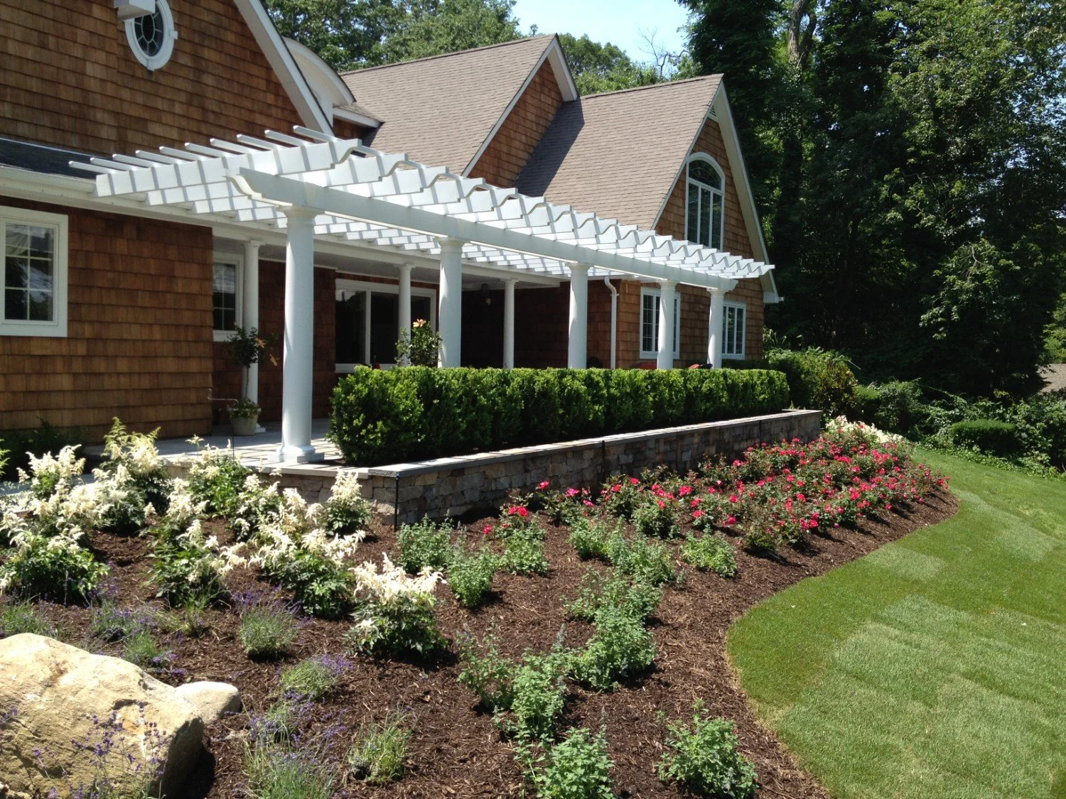 White wood pergola with landscaped garden beds and stone retaining wall at a Setauket, NY home