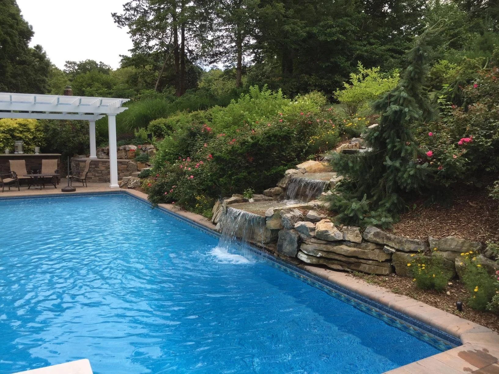 Outdoor living water feature with custom pool, stone waterfall, and surrounding landscape design in Stony Brook, NY