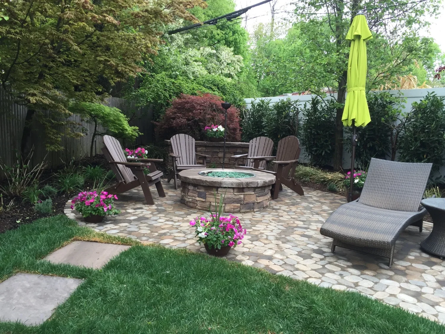 A backyard patio with a fire pit surrounded by five wooden chairs, a lounge chair, and a yellow patio umbrella. There are pink potted flowers, green trees, and shrubs.