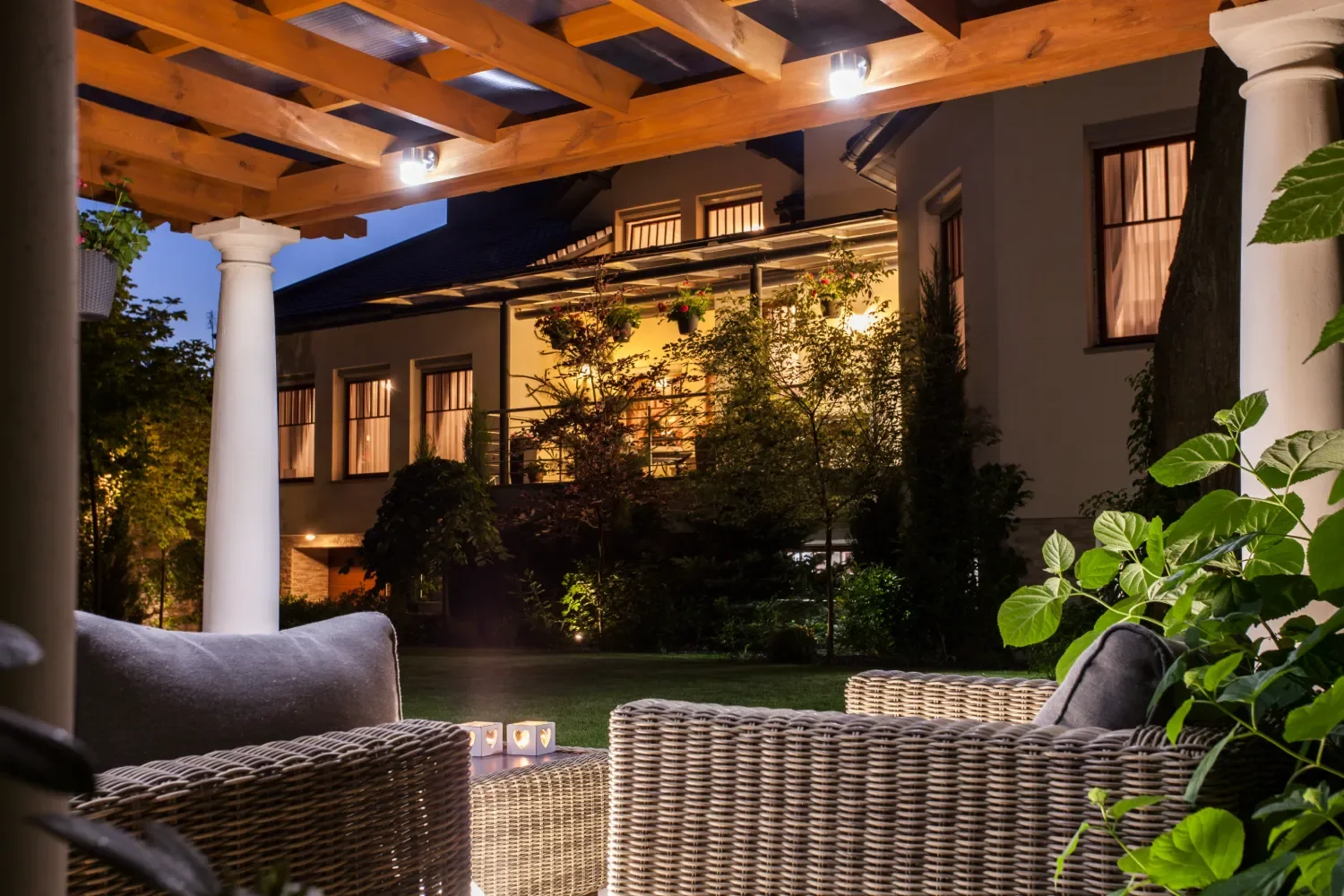 Nighttime view of a cozy backyard patio with wicker chairs, cushions, and lit candles, overlooking a well-lit house with large windows and potted plants under a wooden pergola.