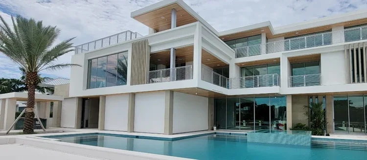 Modern multi-story house with large glass windows, balconies, and a swimming pool in the foreground, with palm trees nearby.