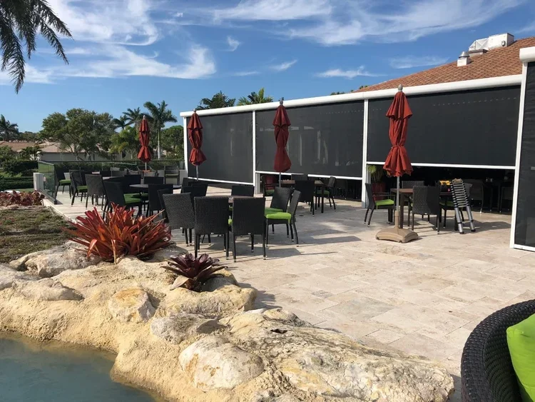 Outdoor patio area with tables, chairs, and red umbrellas, surrounded by rocks, plants, and a pool, with a screened enclosure and house in the background.