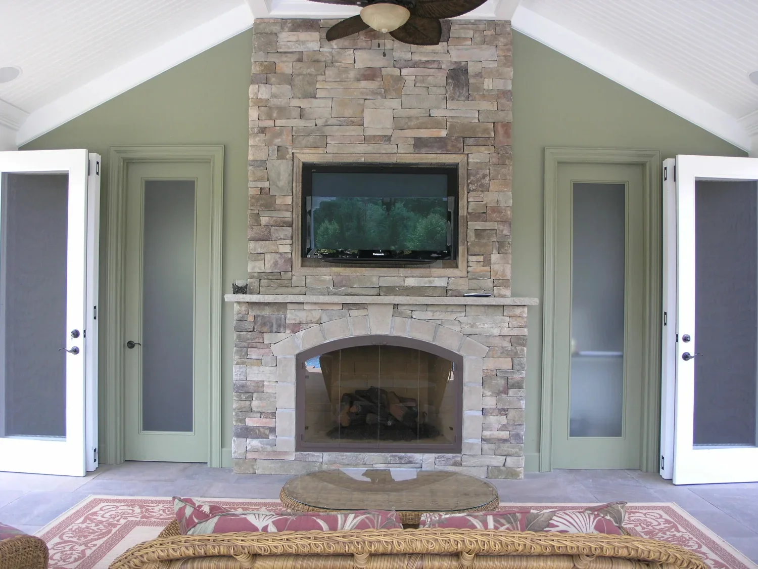 Living room with a stone fireplace, mounted flat-screen TV, green walls, glass doors, ceiling fan, and a round coffee table with a sofa and cushions in the foreground.