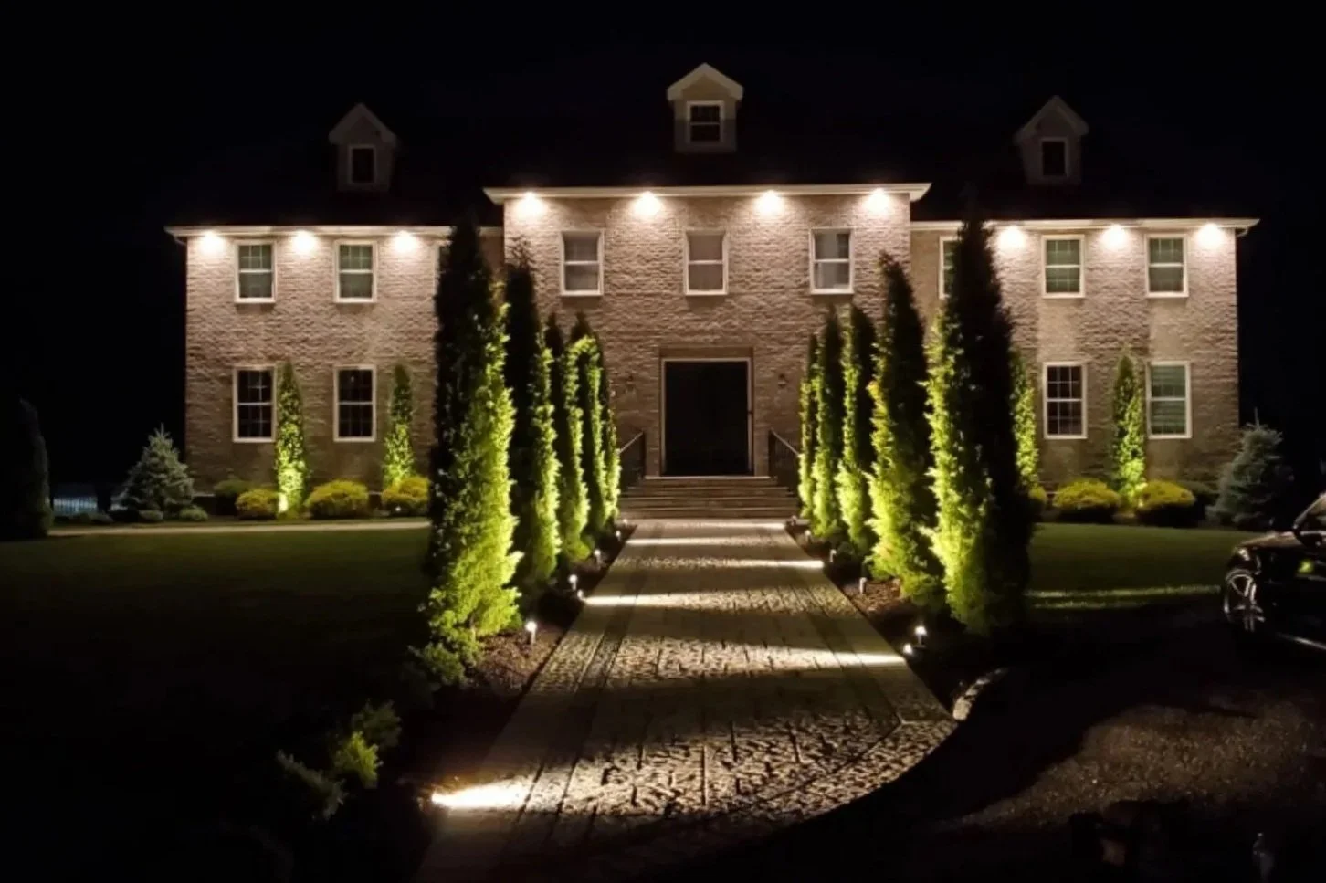 Night view of a large brick house illuminated with exterior lights, with a central walkway leading to the front door flanked by tall trees and bushes.