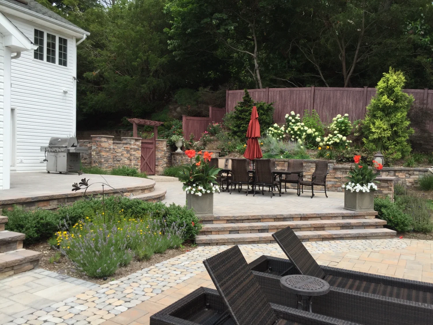 Outdoor living patio and masonry transformation with stone patio, retaining walls, stairs, and layered plantings in Old Westbury, NY