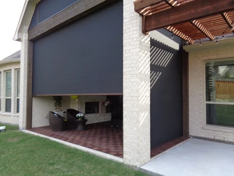 A covered outdoor patio area with sitting furniture and a fireplace, adjacent to a brick house with a large black window shade and a wooden pergola overhead.