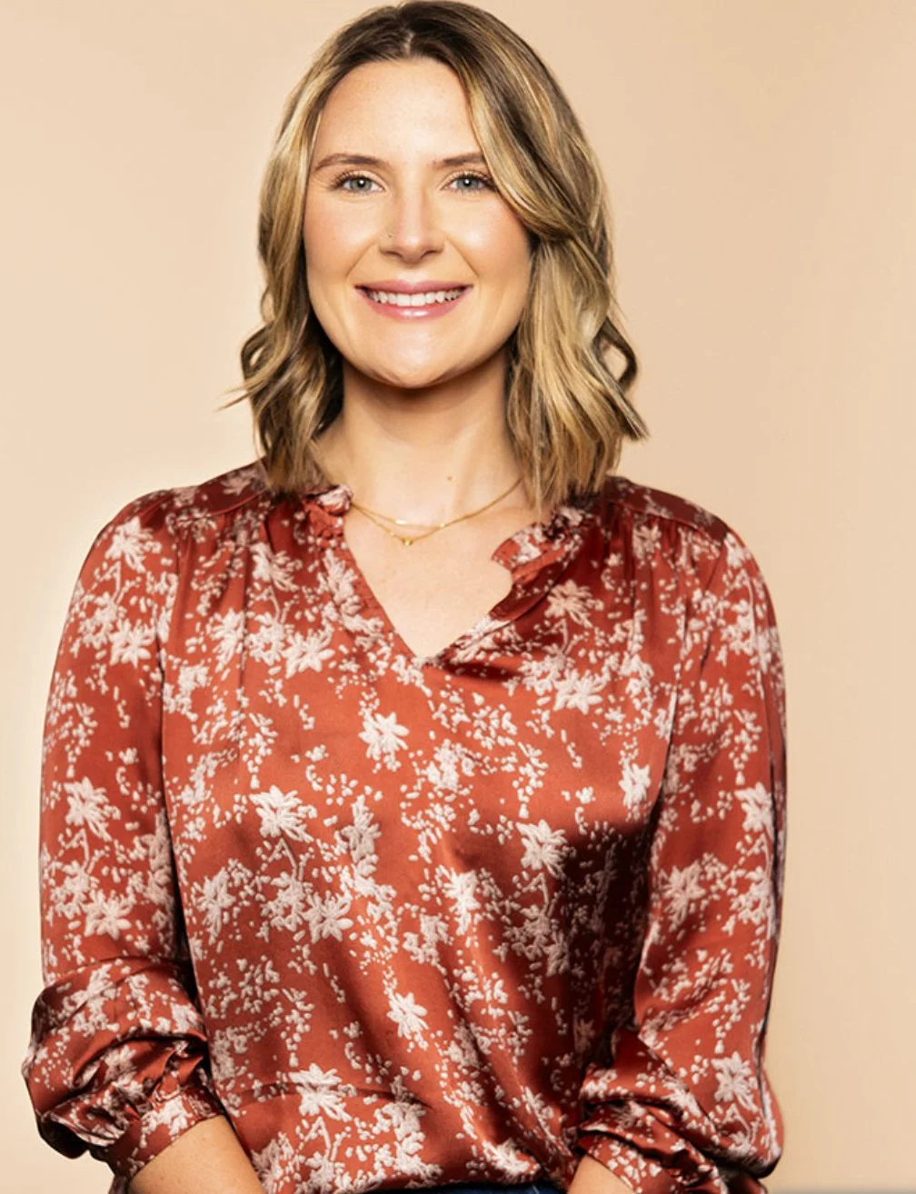 A woman with blonde hair wearing a floral patterned rust-colored blouse and a delicate gold necklace smiling in front of a beige background.