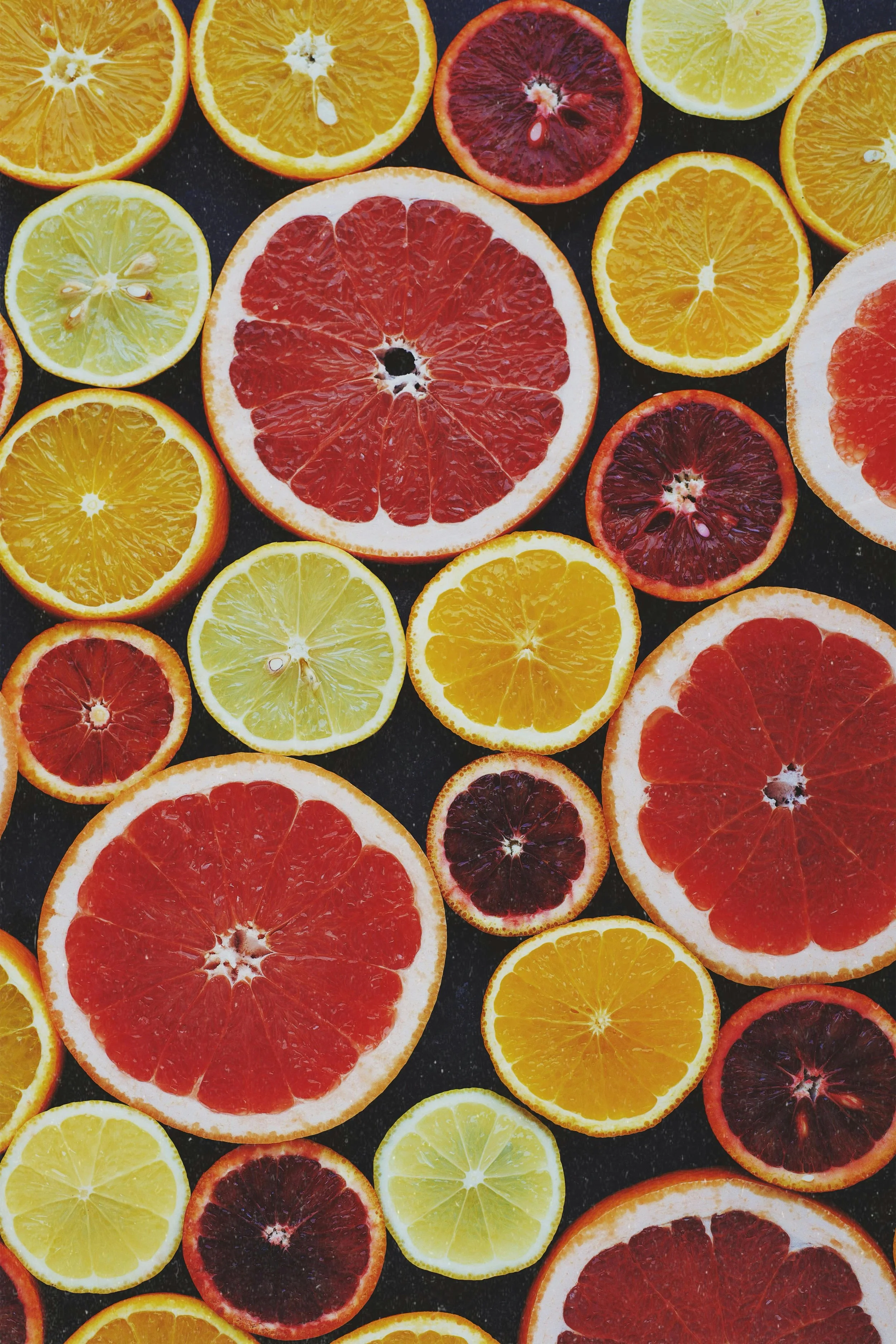A collection of sliced citrus fruits, including grapefruits, oranges, lemons, and blood oranges, arranged on a dark surface.