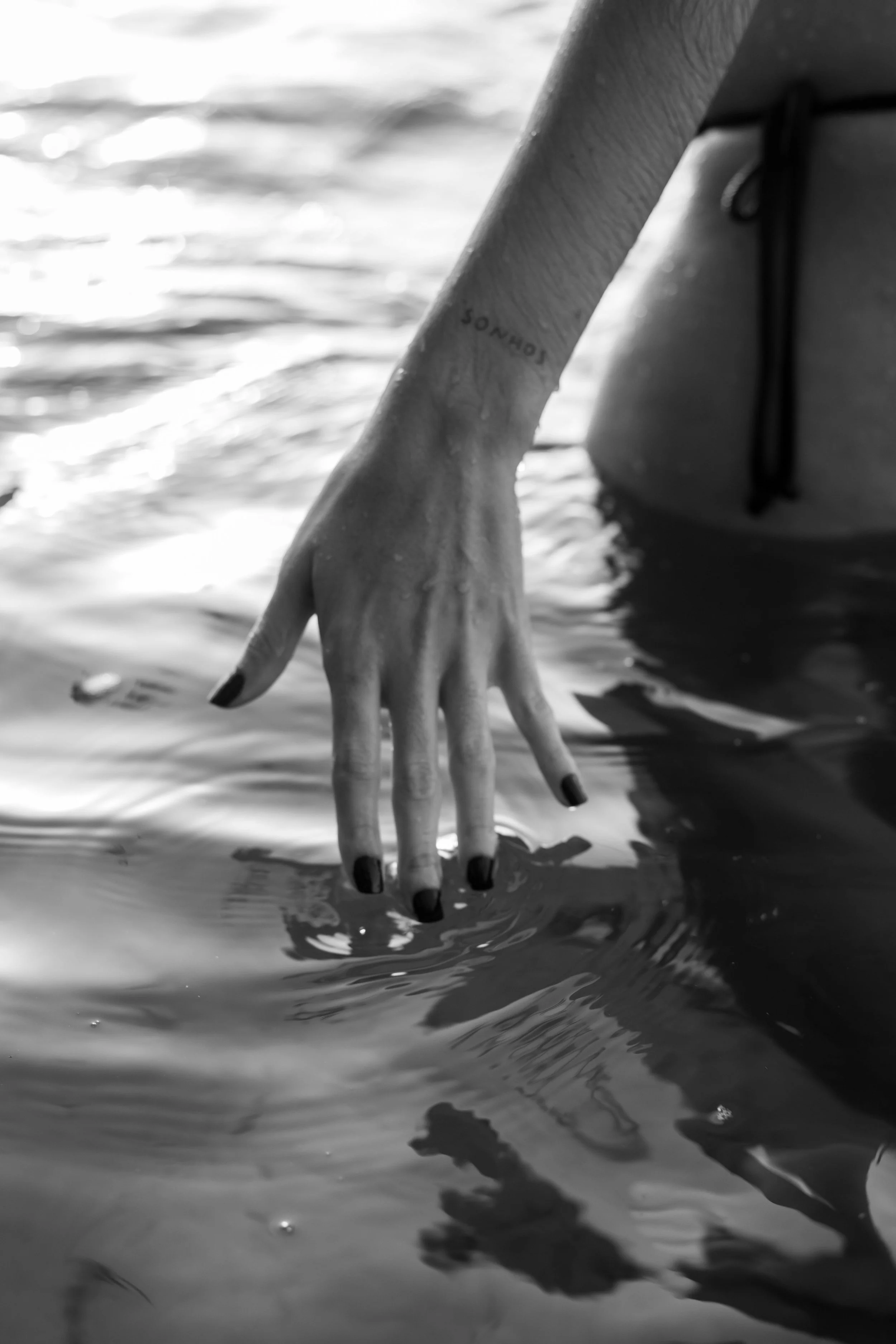 Close-up of a person's hand with black painted nails, dipping into water, with a tattoo on the forearm that says "CONCORD".