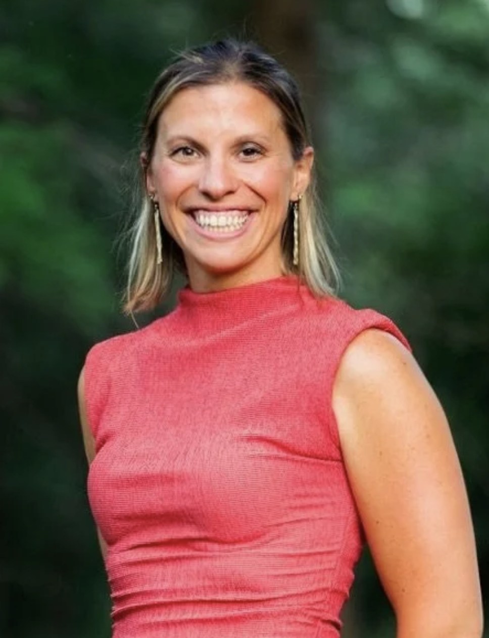 A smiling woman with shoulder-length blonde hair, wearing a sleeveless red top and long earrings, standing outdoors with green blurred foliage in the background.