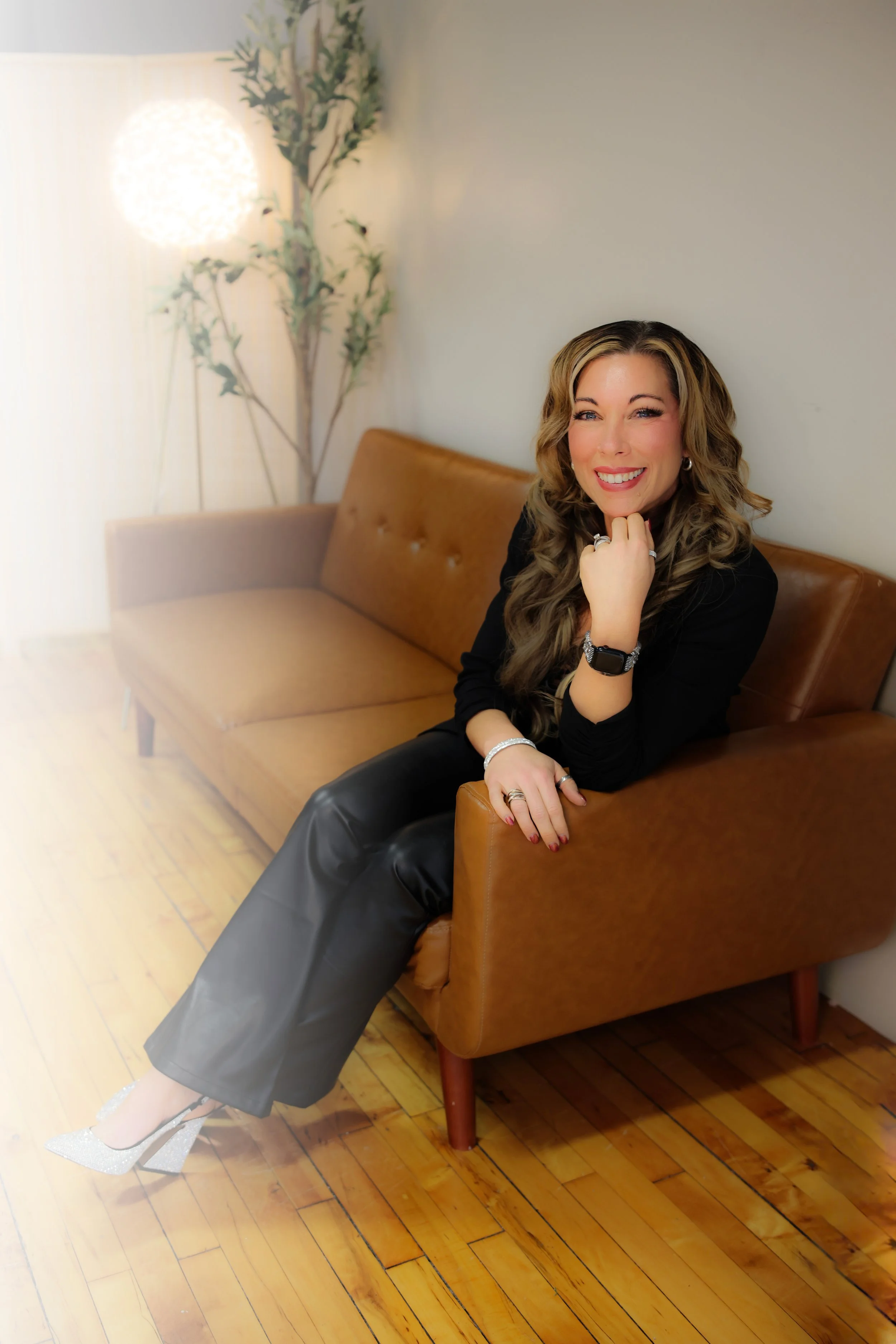 Woman with long wavy hair sitting on a brown leather couch, smiling, with one hand resting on her chin and the other on her leg, dressed in a black top and leather pants, wearing silver and black accessories, in a cozy room with wooden floors, a faux plant, and a glowing lamp in the background.